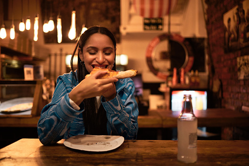 Pizza, food and restaurant where young woman is eating food and enjoying alcohol drink. Hungry female having delicious meal at bar at night. Happy  and satisfied foodie having tasty supper at a diner