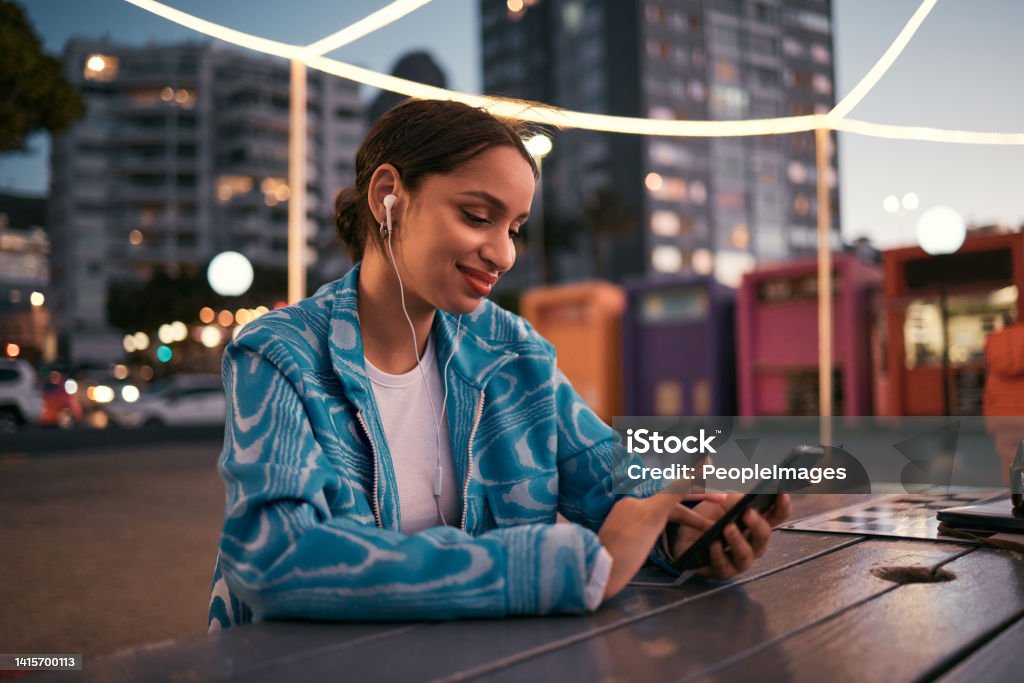 Happy and trendy woman browsing on phone while wearing earphones and listening to music while sitting at outdoor cafe in night city. Happy woman streaming subscription app or making video call Happy, urban and trendy woman browsing on phone while wearing earphones and listening to music while sitting at outdoor cafe in night city. Happy woman streaming subscription app or making video call Mobile Phone Stock Photo
