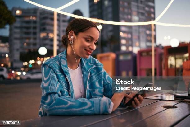 Happy And Trendy Woman Browsing On Phone While Wearing Earphones And Listening To Music While Sitting At Outdoor Cafe In Night City Happy Woman Streaming Subscription App Or Making Video Call Stock Photo - Download Image Now