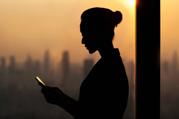 Silhouette of young woman using smartphone next to window with cityscape stock photo