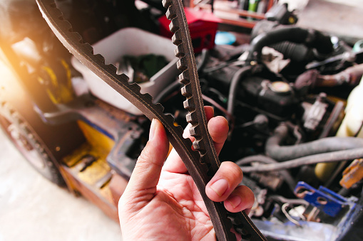A mechanic hand holding a timing belt with a car engine is blurred in the background.