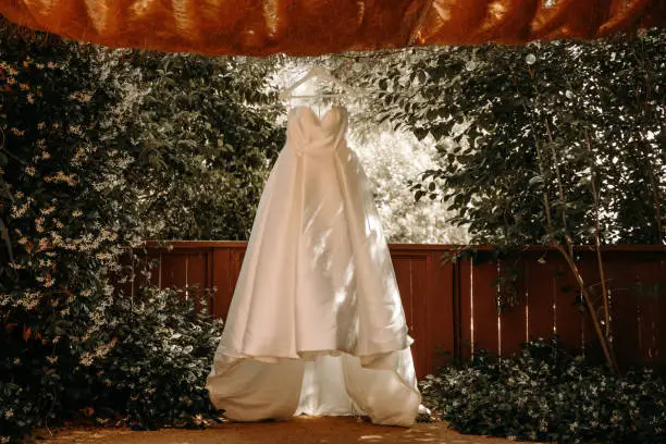 Beautiful wedding dress with pleats and tail hanging on white hanger outdoors. Ballgown in the garden.
