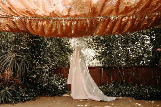 Beautiful wedding dress with pleats and tail hanging on white hanger outdoors. Ballgown in the garden.