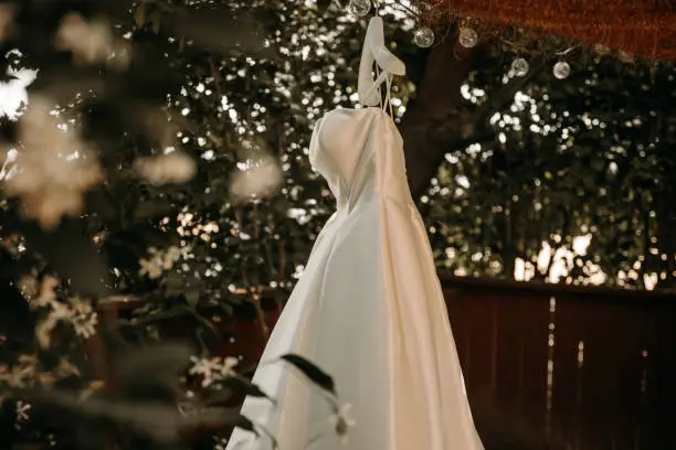 Beautiful wedding dress with pleats and tail hanging on white hanger outdoors. Ballgown in the garden.