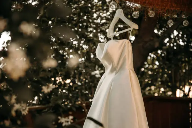 Beautiful wedding dress with pleats and tail hanging on white hanger outdoors. Ballgown in the garden.