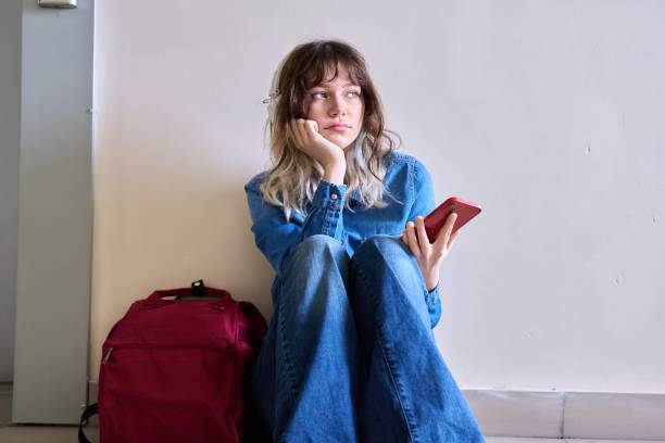 istock Sad young female student with backpack smartphone sitting on the floor 1415714721