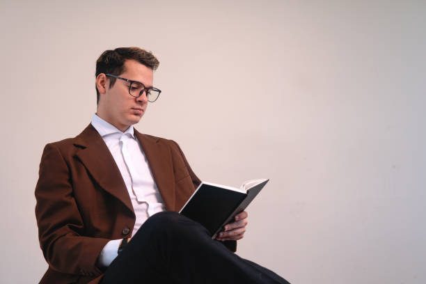 istock Intellectual man dressed in a suit and glasses reading book sitting on a white background. Elegant business man reading his book. University professor with a book in his hands. 1415716025