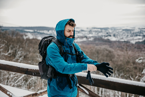 Spontaneous image of a cool looking, bearded, guy, hiking in a snow. He's putting on the snow glows, wearing a blue ski jacket and a hikers backpack. Ready for adventure outdoors on a snow cold day