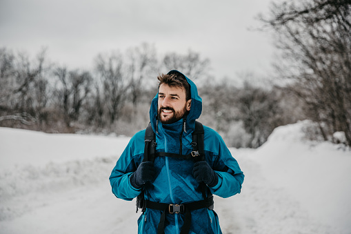 Cool looking, bearded young adult guy, in a ski outfit, enjoying hiking and the snow. He's wearing a ski outfit, with glows and hoodie on, facing away smiling. Radiating active, healthy and positive. Having a backpack, ready for adventure