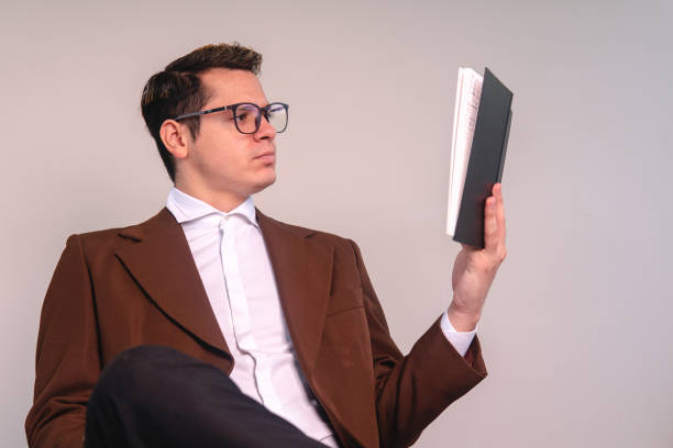 istock A university professor on an isolated white background sitting attentively reading a book. An elegant man studying. Young man dressed in brown suit and wearing glasses. 1415717387