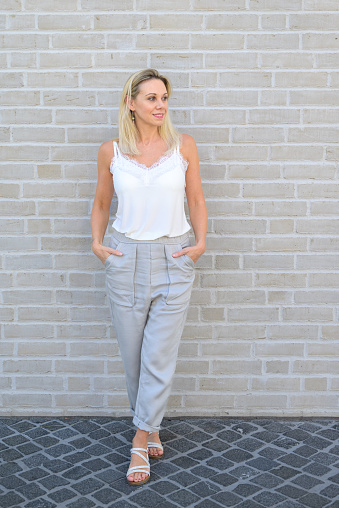 Trendy blond woman posing full length in summer fashion wearing a white top and pants standing against a brick wall