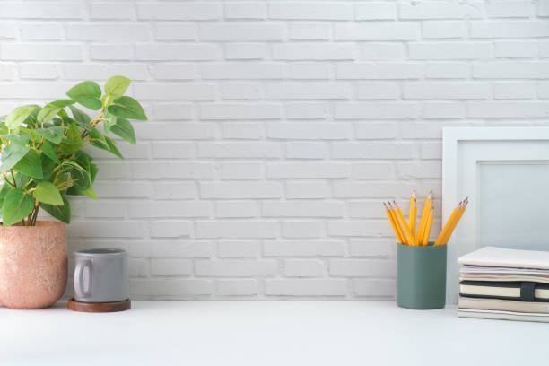 istock Creative workspace with blank poster frame, coffee cup, books and flower pot on white table with brick wall. 1415724025