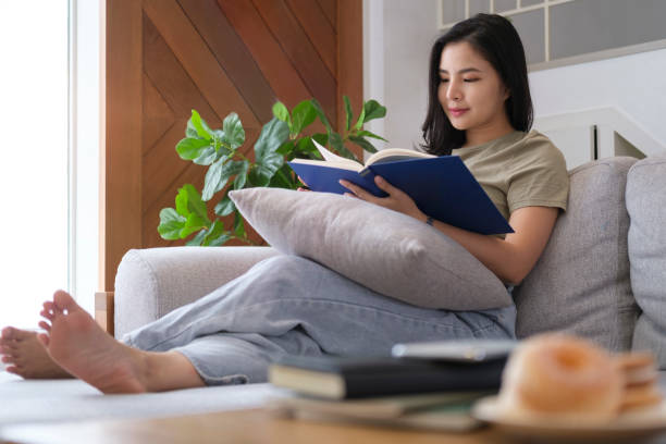 istock Pleased beautiful asian woman reading book on comfortable sofa at home. 1415725607