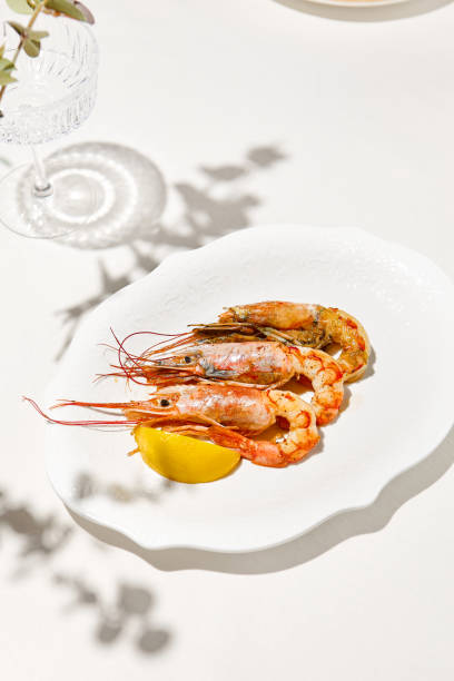 istock Grilled shrimps on light background with shadows. Grilled prawns with lemon on white table. Aesthetic composition with bbq shrimps. Bbq prawns. Grilled seafood. Summer menu. 1415737436