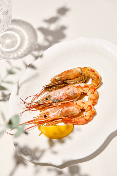istock Grilled shrimps on light background with shadows. Grilled prawns with lemon on white table. Aesthetic composition with bbq shrimps. Bbq prawns. Grilled seafood. Summer menu. 1415737814