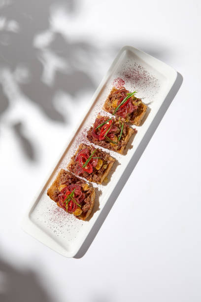 istock Aesthetic composition with shredded beef bruschetta on white background with shadows from flowers. Italian bruschetta with shredded beef and mustard on fine dining in summer. Elegant menu concept. 1415737852