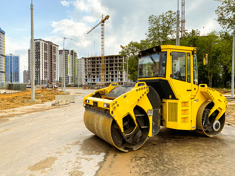 construction machinery builds a large residential area. laying communications. bright, yellow asphalt paver compacts asphalt on the road against the backdrop of tall residential buildings.