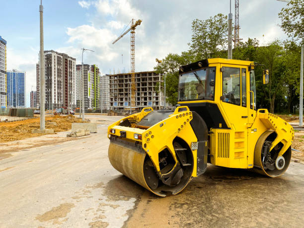 construction machinery builds a large residential area. laying communications. bright, yellow asphalt paver compacts asphalt on the road against the backdrop of tall residential buildings - 工業音樂 個照片及圖片檔