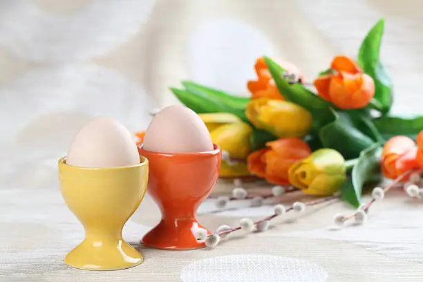 Eggs in orange and yellow eggcups with tulips and catkins in background. 