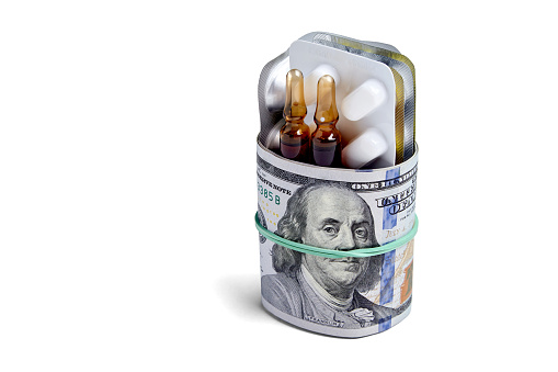 Tablets and ampoules in a blister pack are wrapped in one hundred dollar bills of money, fastened with an elastic band isolated on a white background. Rising drug prices