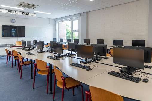 Computer education in classroom on high school. On this long table you see many desktop computers with keyboards and computer mouses. Also many chairs for the children to sit and work with the computer. In this classroom the students get computer lessons so they get prepared for our digital society as a lifestyle