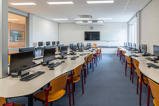 Computer education in classroom on high school. On this long table you see many desktop computers with keyboards and computer mouses. Also many chairs for the children to sit and work with the computer. In this classroom the students get computer lessons so they get prepared for our digital society as a lifestyle