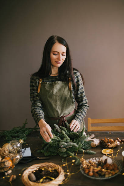 istock Millennial woman making Christmas wreath using natural pine branches and festive decorations in workshop. 1415741806