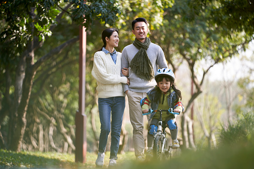 young asian couple taking a walk outdoors in park while daughter riding a kid bike happy and smiling