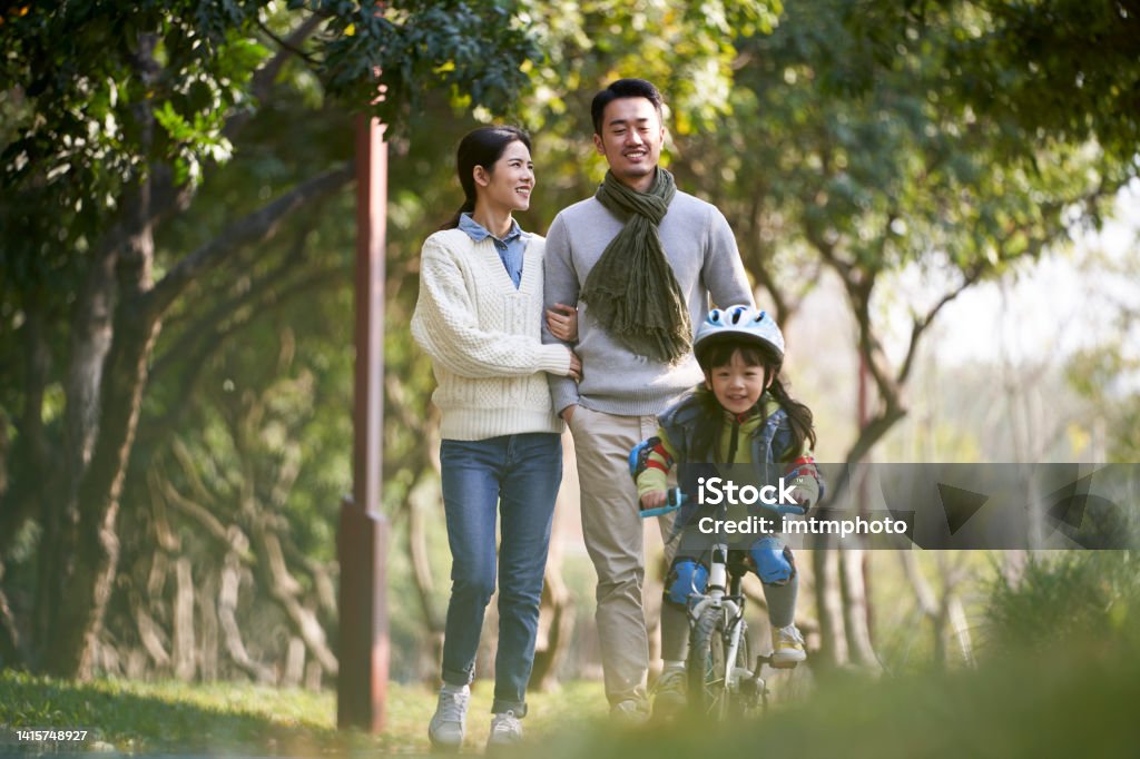 young asian family with one child enjoying outdoor activity in park young asian couple taking a walk outdoors in park while daughter riding a kid bike happy and smiling Family Stock Photo