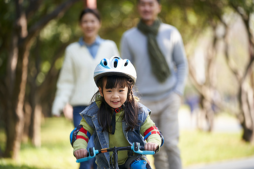 little asian girl with helmet and full protection gears riding bike in city park with parents watching from behind