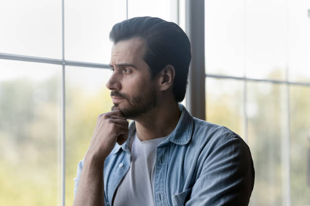 istock Pensive millennial bearded guy stand by window look at distance 1415754389