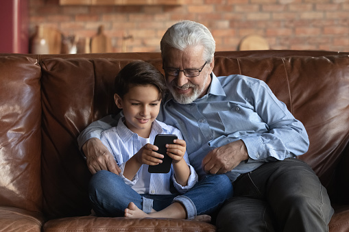 Mutual interests of grandson and grandpa. Happy small grandkid sit on sofa show older grandfather video blog on cell screen. Mature old man have fun with preteen boy watch grandchild play phone game