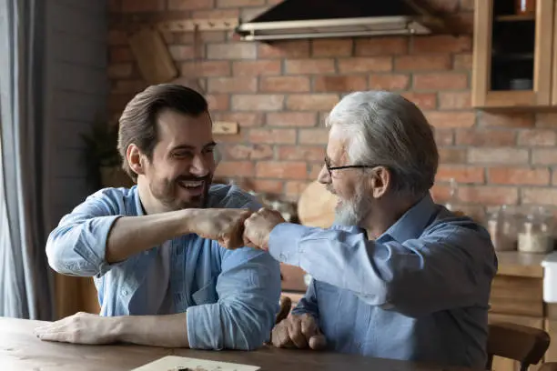 Friendly draw. Happy joyful two men of different age give fist bump after ending game of checkers draughts. Laughing elderly father retiree and millennial son greet each other with finishing boardgame