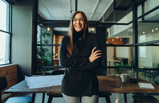 Female business professional smiling at the camera while standing with her arms crossed. Cheerful young businesswoman standing in the boardroom of a modern workplace.