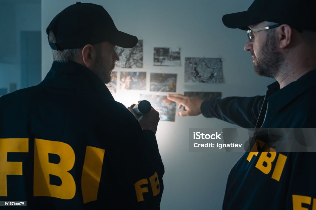 FBI agents at work exploring photos on the wall during investigation Two FBI agents at work. Looking at photos on the wall at victim's apartment, we see them from the waist up from the back. FBI Stock Photo