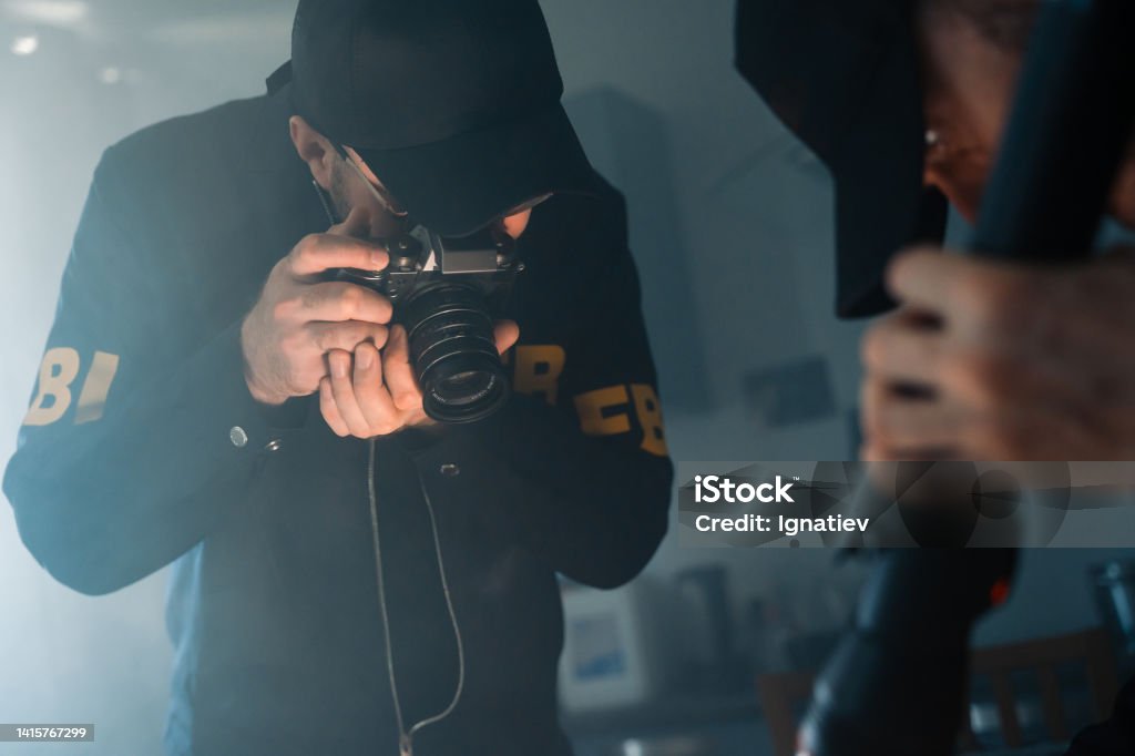 FBI agents at the crime scene taking pictures of physical evidences on a table in a flashlight light FBI agents in uniform taking pictures of physical evidences on a table with camera at the den of criminals in a dark room. FBI agents at the crime scene FBI Stock Photo
