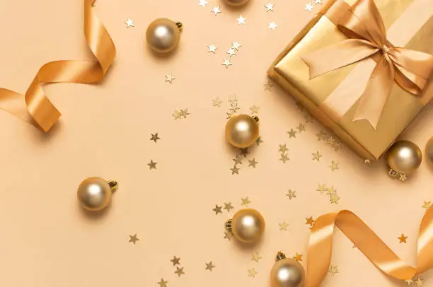 Photo of Merry Christmas and Happy Holidays greeting card. Beautiful golden gift balls ribbons confetti stars on gold background top view Flat lay. New Year presents Festive decorations 2020 celebration