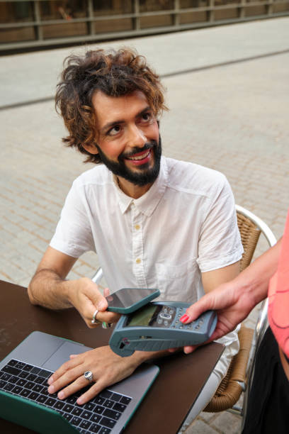 istock Man with makeup smiling while making a contactless payment with a mobile phone in an outdoor cafe. 1415778138