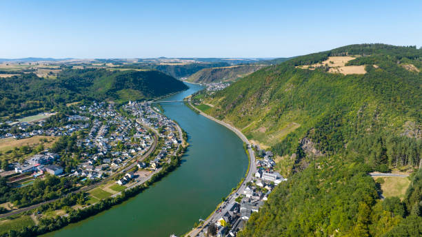 A scenic aerial view of a barge navigating the winding Moselle River near the steep Calmont vineyards. The lush green valley, terraced vineyards, and charming village in the distance showcase the beauty of Germany's wine country.