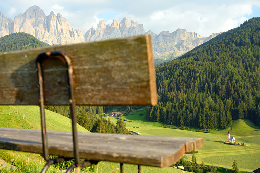 (Selective focus) Defocused park bench in the foreground with the Church of St. John (San Giovanni in Ranui) that stands out in the green meadows, in the heart of the beautiful Dolomitic mountain landscape.