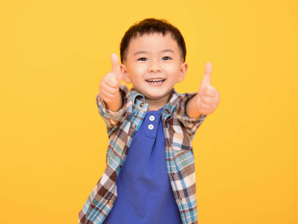 黄色の背景の前で幸せな小さな男の子と親指を上に表示 - child facial expression asian and indian ethnicities asia ストックフォトと画像