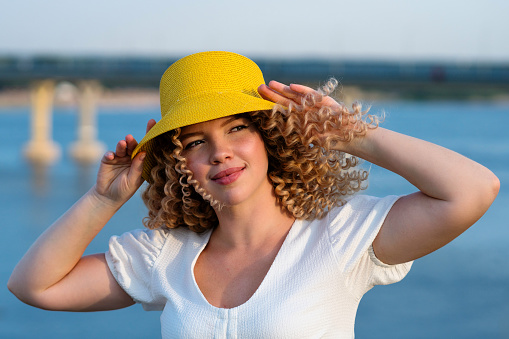 Glad romantic girl in yellow bucket hat panama outdoor looks away. Young caucasian woman with afro curls hairstyle at sunset in summer sea background in windy day