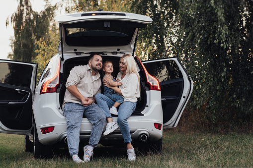 Young Family Sitting Inside Opened Trunk of SUV Car, Happy Parents with Their Little Daughter Enjoying Road Trip
