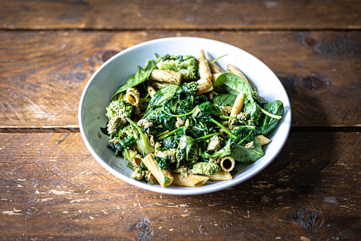A picture from above of a white plate of whole-wheat pasta with spinach and broccoli on a wooden table.
