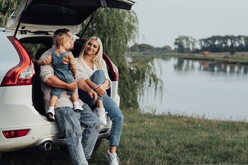 Young Parents with Little Daughter Sitting in Open Trunk of Car, Happy Family Enjoying Road Trip on Their SUV Car