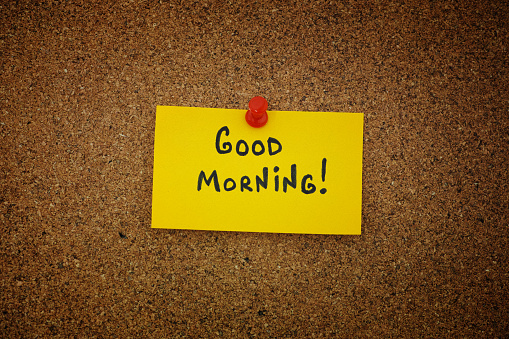 A yellow paper note with with the phrase Good Morning! on it pinned to a cork board. Close up.