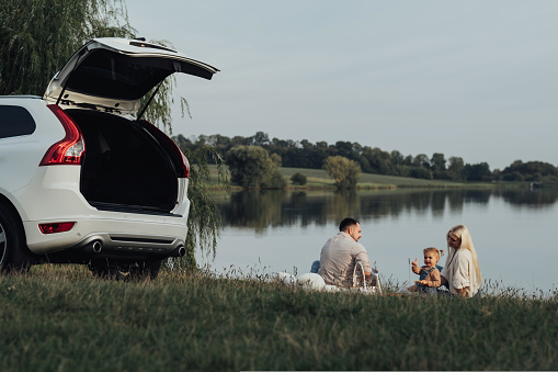 Happy Young Family Enjoying Picnic Time Outside City, Mother and Father with Their Daughter Sitting Outdoors Near Their SUV Car, Road Trip Concept