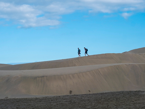 Maspalomas, Gran Canaria, Canary Islands, Spain December 18, 2020: Two barefoot man couple walking at the edge of sand dune at Natural Reserve of Dunes of Maspalomas, blue sky background.
