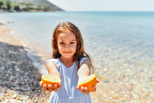 Happy little girl on the beach playing and eating orange and enjoying it.