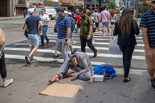 6th Avenue, Manhattan, New York, NY, USA - July 8th 2022: Man surrounded by pedestrians sitting on the sidewalk begging for money